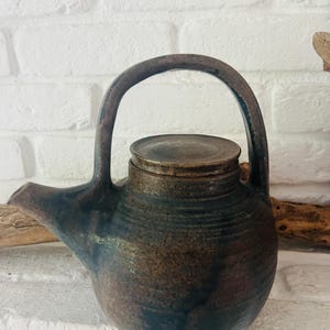 Könnte beinhalten: Eine handgefertigte Keramik-Teekanne mit dunkler, gesprenkelter Glasur in Braun- und Blautönen. Die Teekanne hat einen abgerundeten Körper, einen gebogenen Griff, einen Ausguss und einen Deckel. Es ist ein einzigartiges Keramikstück.