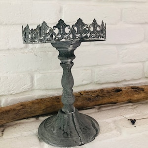 Könnte beinhalten: Ein grauer Kuchenständer aus Metall mit einem dekorativen, verzierten Oberteil. Der Ständer hat einen hohen, schlanken Fuß und eine runde Oberseite.