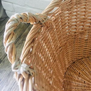 May include: A large, natural woven wicker basket with a single handle. The basket is made of light brown wicker and has a natural, rustic look.