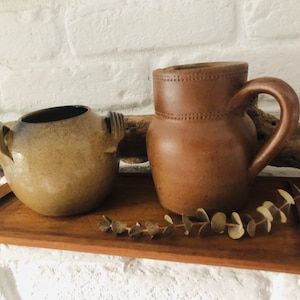 May include: Two brown ceramic pottery pieces, a pitcher with a handle and a round pot with two handles, are displayed on a wooden tray with a sprig of dried eucalyptus.