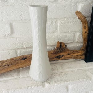 Könnte beinhalten: Eine weiße Keramikvase mit einem strukturierten Design. Die Vase ist hoch und schlank, mit einer schmalen Öffnung an der Oberseite.