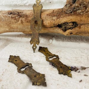 Könnte beinhalten: Drei antike Messingbeschläge, darunter eine Schlüssellochabdeckung und zwei dekorative Platten, werden vor einer weißen Backsteinmauer und einem Stück Treibholz präsentiert. Die Beschläge haben ein gealtertes, angelaufenes Aussehen, was auf Vintage- oder Recycling-Ursprünge hindeutet.