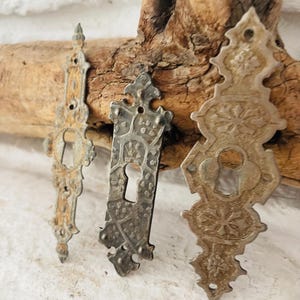 Peut inclure: Trois caches-trous de serrure antiques en métal aux motifs ornés. Les caches sont dans des tons argentés et bruns variés, montrant des signes d'âge et d'usure. Les caches-trous de serrure sont disposés sur un morceau de bois vieilli sur fond blanc.