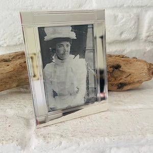 Könnte beinhalten: Ein rechteckiger silberner Bilderrahmen mit einem Schwarz-Weiß-Foto einer Frau mit Hut und Kleid. Der Rahmen hat ein schlichtes, elegantes Design und steht auf einer weißen Oberfläche.