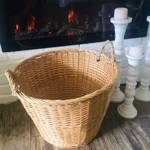 May include: A large, natural woven wicker basket with two rope handles. The basket is sitting on a wooden floor in front of a fireplace with white candlesticks.