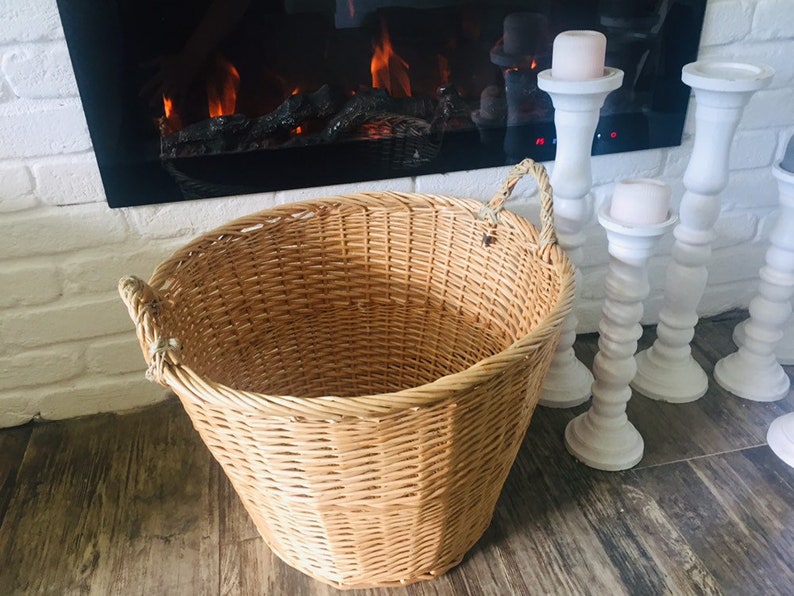 May include: A large, natural woven wicker basket with two rope handles. The basket is sitting on a wooden floor in front of a fireplace with white candlesticks.