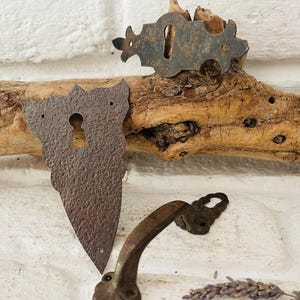 Könnte beinhalten: Nahaufnahme von antiken Metallbeschlagteilen, darunter eine Schlüssellochabdeckung, eine Rosette und ein Griff. Die Teile haben ein rustikales, gealtertes Aussehen mit dunkler, verwitterter Oberfläche. Die Hardware wird vor einem weißen Ziegelhintergrund präsentiert.