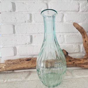 Könnte beinhalten: Eine hohe, transparente Glasvase mit leichtem Türkis-Ton. Die Vase hat ein geripptes Design und einen ausgestellten oberen Rand. Der Hintergrund zeigt eine weiße Backsteinmauer und ein Stück Treibholz.