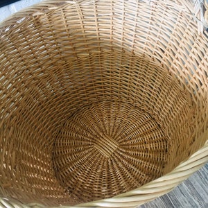 May include: A large, round wicker basket with a woven handle. The basket is made of natural, light brown wicker and has a white rim.