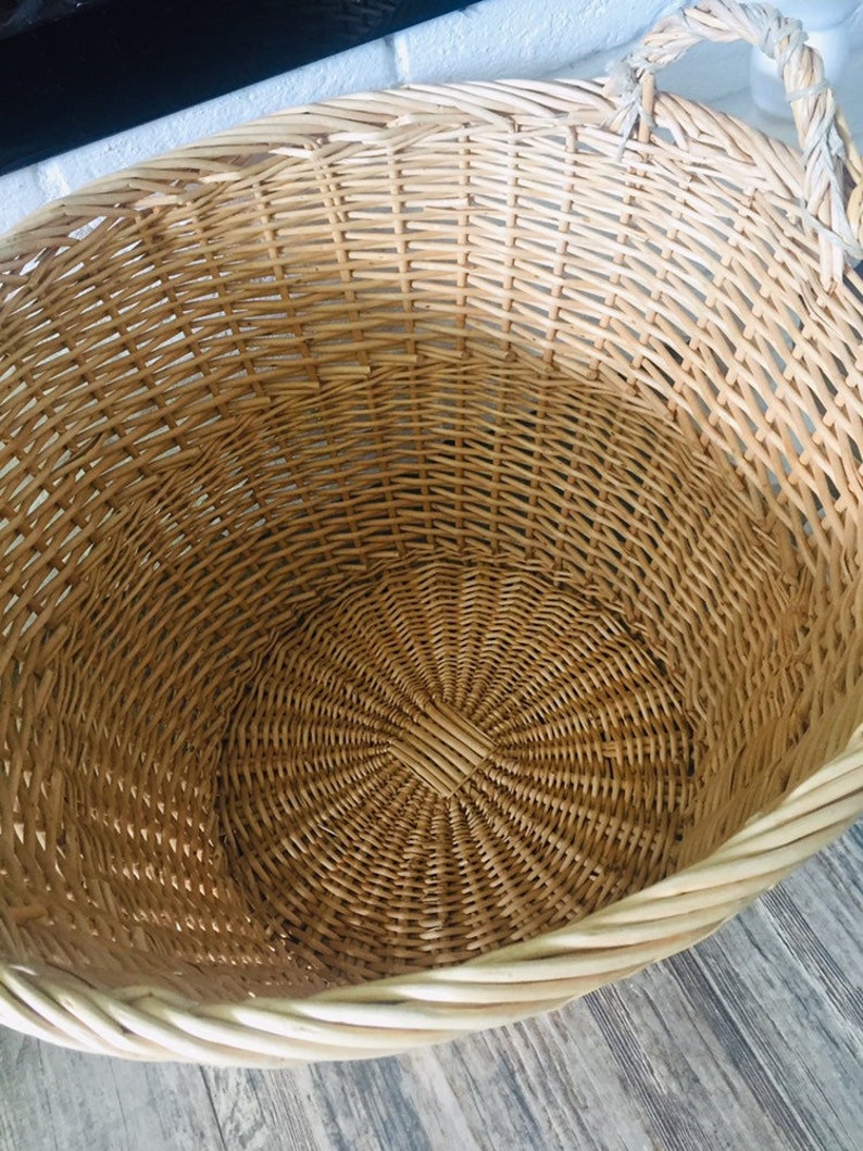 May include: A large, round wicker basket with a woven handle. The basket is made of natural, light brown wicker and has a white rim.
