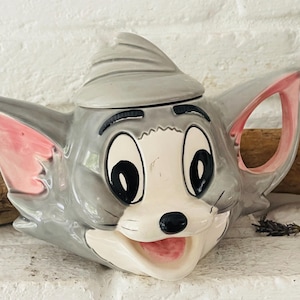 Könnte beinhalten: Eine Keramik-Teekanne in Form eines Cartoon-Katzenkopfes, grau und weiß mit rosa Akzenten in Ohren und Mund. Die Teekanne hat einen Deckel und einen Henkel und steht vor einem weißen Ziegelhintergrund.