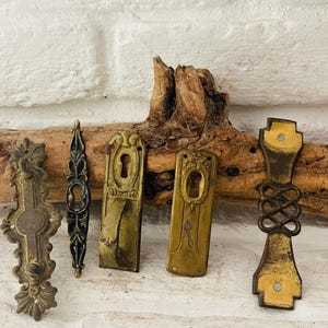 May include: An assortment of antique brass door hardware, including ornate handles and keyhole covers, arranged on a white brick surface. The hardware pieces vary in design, showcasing intricate patterns and aged patina. A sprig of lavender adds a touch of natural contrast.
