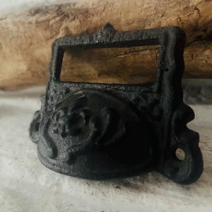 Könnte beinhalten: Ein schwarzer, verzierter Schubladengriff aus Gusseisen mit einem rechteckigen Griff und einem dekorativen Blumendesign. Die Hardware hat ein strukturiertes, gealtertes Aussehen, das einen Vintage-Stil suggeriert. Ideal für Möbelrestaurierung.