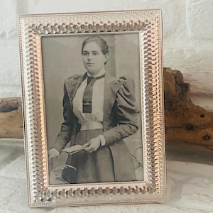 Könnte beinhalten: Rechteckiger silberfarbener Bilderrahmen mit strukturiertem Rand. Der Rahmen enthält ein Schwarz-Weiß-Porträt einer Person in Vintage-Kleidung. Das Porträt befindet sich vor einer weißen Backsteinmauer.