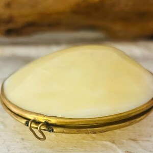 May include: A cream-colored, clam-shaped trinket box with a brass-colored metal frame and clasp. The box has a smooth, rounded top and a hinged opening. The background is a blurred mix of textures and colors.