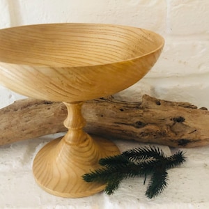 Könnte beinhalten: Eine Holzschale mit einem Sockel. Die Schale ist aus hellem Holz mit natürlicher Maserung. Die Schale ist leer und bereit, mit Obst, Blumen oder anderen Dekorationsgegenständen gefüllt zu werden.