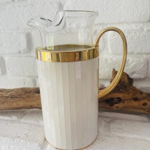 Könnte beinhalten: Eine weiß-goldene Kanne mit einem Ausguss aus klarem Glas und einem goldenen Griff. Die Kanne hat vertikale weiße und hellbeige Streifen. Der Griff ist aus gebogenem Goldmetall. Die Kanne steht auf einer weißen Oberfläche.