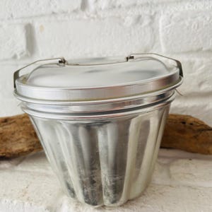 Könnte beinhalten: Eine geriffelte Aluminiumform mit Deckel. Die Form hat eine abgerundete Form mit vertikalen Rippen und einem Metallgriff. Der Deckel wird mit einem Metallverschluss befestigt. Die Form wird wahrscheinlich zum Backen oder zur Herstellung von Desserts verwendet.