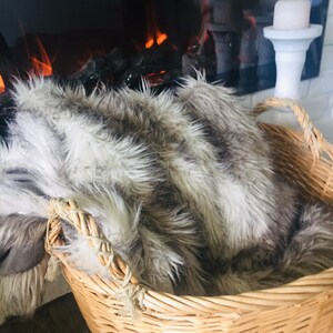 May include: A wicker basket with a faux fur throw inside. The basket has two handles and is sitting in front of a fireplace.