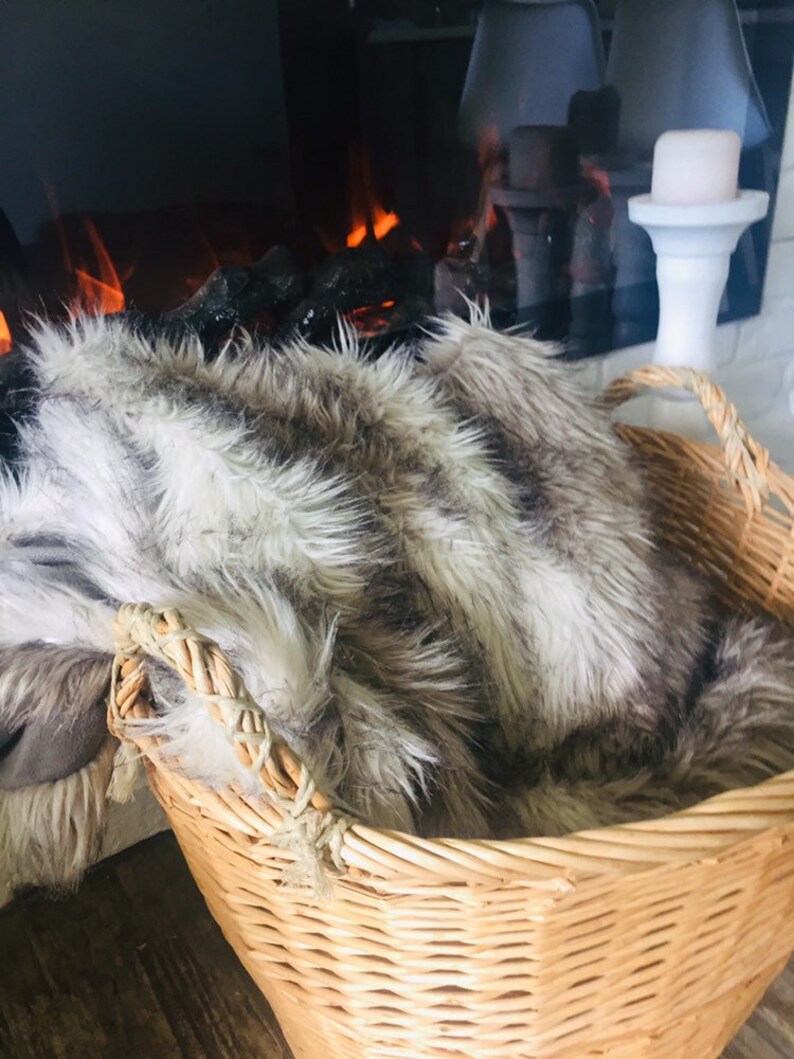 May include: A wicker basket with a faux fur throw inside. The basket has two handles and is sitting in front of a fireplace.