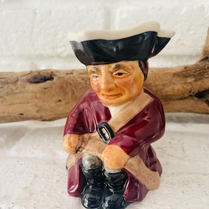Könnte beinhalten: Keramikfigur eines Mannes mit Dreispitz und burgunderfarbenem Mantel. Die Figur hat ein hellbraunes Gesicht und Hände, schwarze Stiefel und eine beige Schärpe. Die Figur sitzt, mit weißem Innenraum.