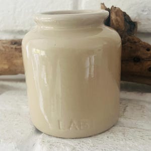 Könnte beinhalten: Ein kleiner, beigefarbener Keramiktopf mit zylindrischem Körper und leicht ausgestelltem Rand. Das Wort "LAB" ist auf der Vorderseite eingeprägt. Der Topf hat eine glatte, glänzende Oberfläche und scheint Vintage oder antik zu sein. Der Hintergrund umfasst eine weiße Ziegelmauer und ein Stück Treibholz.