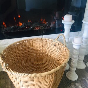 May include: A large, natural woven wicker basket with two rope handles. The basket is empty and sitting on a wood floor in front of a fireplace.
