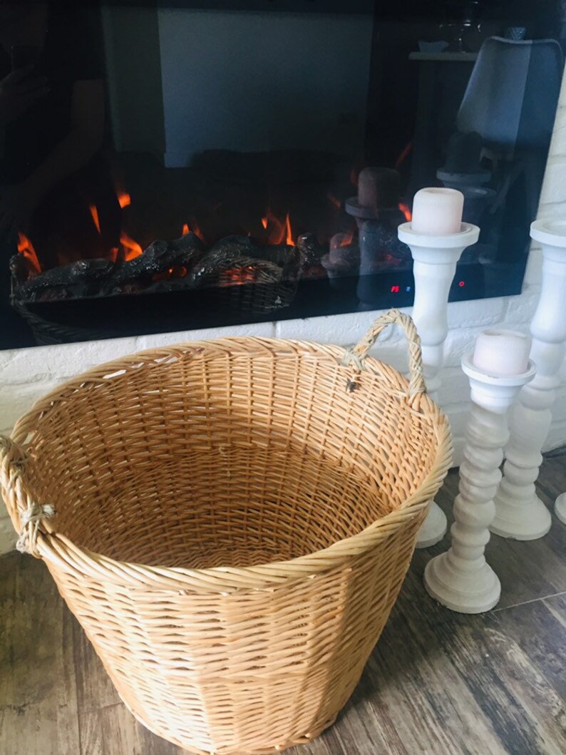 May include: A large, natural woven wicker basket with two rope handles. The basket is empty and sitting on a wood floor in front of a fireplace.