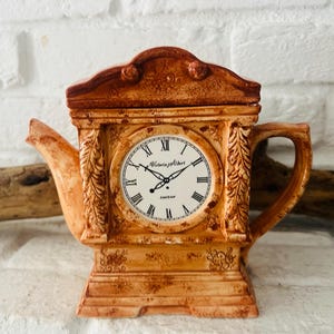 Könnte beinhalten: Eine dekorative Teekanne im Uhr-Design. Die hellbraune Teekanne hat ein Zifferblatt mit römischen Ziffern und den Worten "Victoria & Albert London". Die Teekanne hat einen Griff, einen Ausguss und einen Deckel. Die Zeiger zeigen die Uhrzeit an.