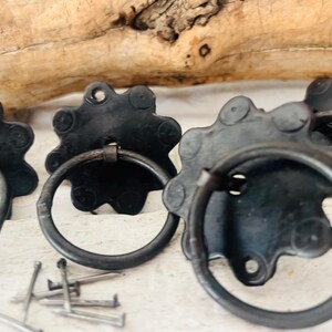 Könnte beinhalten: Nahaufnahme mehrerer schwarzer Metall-Schubladengriffe mit Ringgriffen. Die Griffe haben ein Sternmuster und werden von kleinen Nägeln begleitet. Die Beschläge sind wahrscheinlich für die Möbelrestaurierung oder ein rustikales Dekorationsprojekt gedacht.