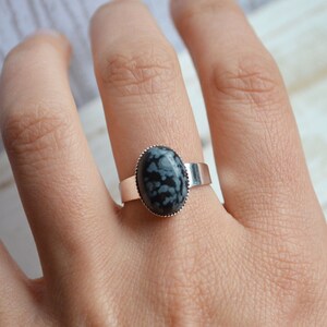 Könnte beinhalten: Ein silberner Ring mit einem schwarzen und weißen Schneeflocken-Obsidian-Stein. Der Ring befindet sich an einem Finger.