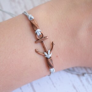 Könnte beinhalten: Ein graues geflochtenes Armband mit einem Anker-Anhänger aus Kupfer. Das Armband ist mit einem Knoten gebunden und hat an jedem Ende eine Perle aus Kupfer.