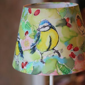 Könnte beinhalten: Konischer Lampenschirm mit einem Aquarelldesign, das gelbe und blaue Vögel, rote Beeren und grüne Blätter zeigt. Der Lampenschirm steht vor einem unscharfen Hintergrund.