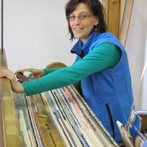 Könnte beinhalten: Eine Frau bedient einen Webstuhl und webt einen bunten Teppich mit verschiedenen Blautönen, Rot, Gelb und Weiß. Der Webstuhl ist aus Holz und hat eine vertikal verlaufende Metallstange. Die Frau trägt eine blaue Weste über einem grünen Hemd.