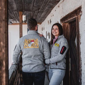 Könnte beinhalten: Zwei Personen tragen graue Fleecejacken mit gestickten Designs. Die Jacke links hat ein Wappen-Design. Die Jacke rechts hat einen kleinen, bunten Aufnäher.