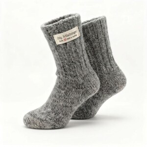 Könnte beinhalten: Ein Paar graue, grob gestrickte Socken mit Rippenstruktur und einem weißen Stoffetikett am Bündchen. Das Etikett zeigt den Text 'Orig. Schladminger Socke, made in Austria' zusammen mit einem kleinen rot-weißen Flaggensymbol.