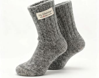 Könnte beinhalten: Ein Paar graue, grob gestrickte Socken mit Rippenstruktur und einem weißen Stoffetikett am Bündchen. Das Etikett zeigt den Text Orig. Schladminger Socke, made in Austria, zusammen mit einem kleinen rot-weißen Flaggensymbol.