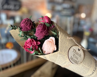 Könnte beinhalten: Ein Strauß aus rosa, burgunderroten und pfirsichfarbenen Sola-Holzblumen, verpackt in braunes Papier mit einer Kordelschleife. Ein rundes Holzschild mit dem Text "Danke Mama!" ist an dem Strauß befestigt.