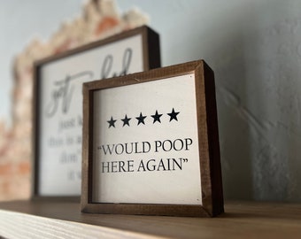 Könnte beinhalten: Ein kleines, quadratisches Holzschild mit dem Text "WOULD POOP HERE AGAIN" in Schwarz, mit fünf schwarzen Sternen über dem Text. Das Schild hat einen dunkelbraunen Rahmen und wird auf einem Holzregal ausgestellt. Ein weiteres Schild ist im Hintergrund zu sehen.