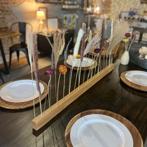 K&ouml;nnte beinhalten: Ein Holzst&auml;nder mit getrockneten Blumen und Gr&auml;sern, die in einer Reihe angeordnet sind. Der St&auml;nder steht auf einem dunklen Holztisch mit zwei wei&szlig;en Tellern und Holztellern.