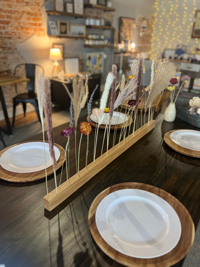 K&ouml;nnte beinhalten: Ein Holzst&auml;nder mit getrockneten Blumen und Gr&auml;sern, die in einer Reihe angeordnet sind. Der St&auml;nder steht auf einem dunklen Holztisch mit zwei wei&szlig;en Tellern und Holztellern.