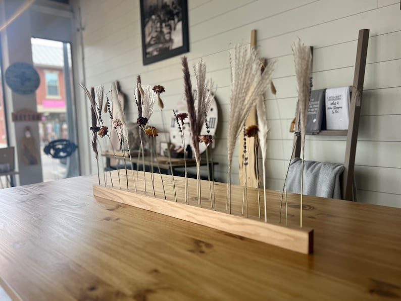 K&ouml;nnte beinhalten: Ein Holzst&auml;nder mit getrockneten Blumen und Gr&auml;sern, die in einer Reihe angeordnet sind. Der St&auml;nder ist aus hellbraunem Holz gefertigt und hat eine nat&uuml;rliche Oberfl&auml;che. Die Blumen und Gr&auml;ser sind in verschiedenen Farben, darunter Wei&szlig;, Braun und Beige.
