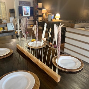 K&ouml;nnte beinhalten: Ein Holztisch mit einer Holztischdecke und wei&szlig;en Tellern. Ein Holz-Mittelst&uuml;ck mit getrockneten Blumen und Gr&auml;sern steht in der Mitte des Tisches.