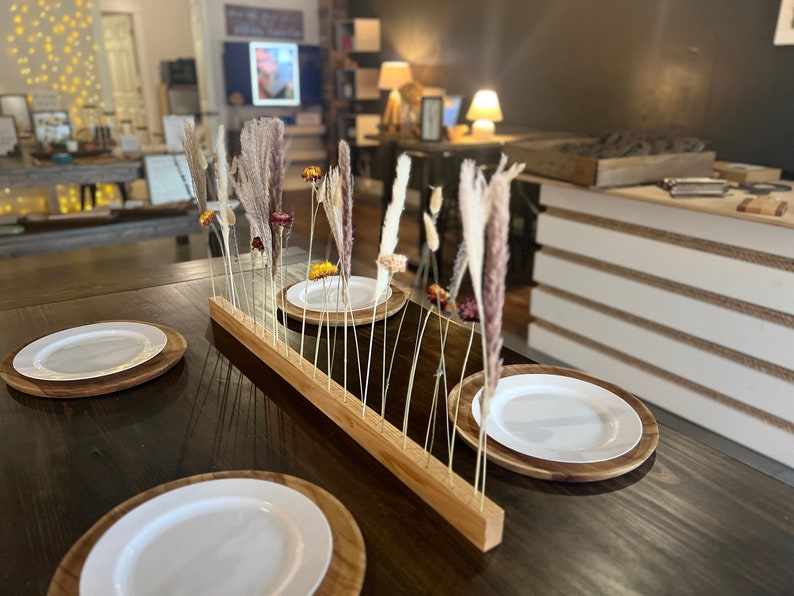 K&ouml;nnte beinhalten: Ein Holztisch mit einer Holztischdecke und wei&szlig;en Tellern. Ein Holz-Mittelst&uuml;ck mit getrockneten Blumen und Gr&auml;sern steht in der Mitte des Tisches.