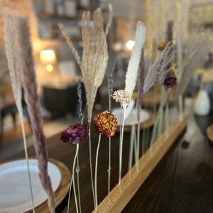 K&ouml;nnte beinhalten: Ein Holzsockel mit getrockneten Blumen und Gr&auml;sern in einer Reihe angeordnet. Die Blumen sind in Braun-, Lila- und Wei&szlig;t&ouml;nen gehalten. Der Sockel steht auf einem dunklen Holztisch.