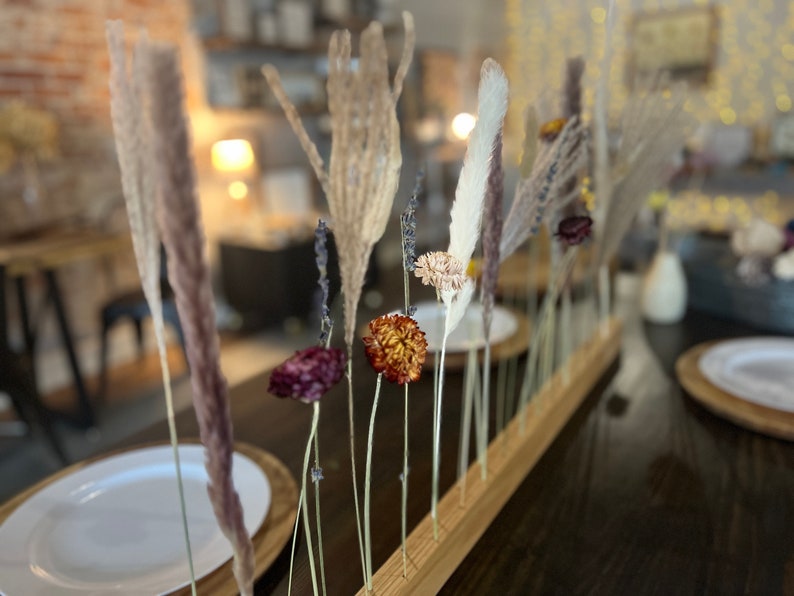 K&ouml;nnte beinhalten: Ein Holzsockel mit getrockneten Blumen und Gr&auml;sern in einer Reihe angeordnet. Die Blumen sind in Braun-, Lila- und Wei&szlig;t&ouml;nen gehalten. Der Sockel steht auf einem dunklen Holztisch.