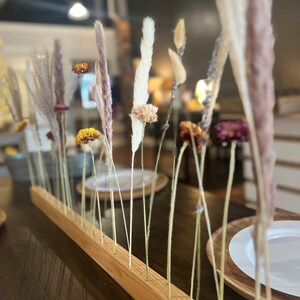 K&ouml;nnte beinhalten: Ein Holzsockel mit L&ouml;chern f&uuml;r getrocknete Blumen und Gr&auml;ser. Der Sockel ist aus hellbraunem Holz und hat eine nat&uuml;rliche Oberfl&auml;che. Die Blumen und Gr&auml;ser sind in verschiedenen Farben, darunter Wei&szlig;, Braun und Lila.