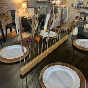 K&ouml;nnte beinhalten: Ein Holz-Tisch-Mittelst&uuml;ck mit getrockneten Blumen und Gr&auml;sern. Das Mittelst&uuml;ck besteht aus einem langen, rechteckigen St&uuml;ck Holz mit gebohrten L&ouml;chern. Die Blumen und Gr&auml;ser sind in den L&ouml;chern angeordnet und haben alle unterschiedliche Farben und Texturen. Das Mittelst&uuml;ck steht auf einem dunkelbraunen Holztisch, und es befinden sich zwei wei&szlig;e Teller auf dem Tisch.