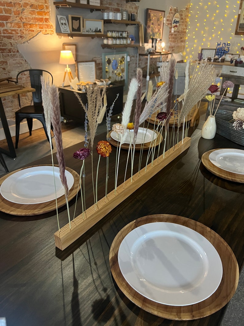 K&ouml;nnte beinhalten: Ein Holz-Tisch-Mittelst&uuml;ck mit getrockneten Blumen und Gr&auml;sern. Das Mittelst&uuml;ck besteht aus einem langen, rechteckigen St&uuml;ck Holz mit gebohrten L&ouml;chern. Die Blumen und Gr&auml;ser sind in den L&ouml;chern angeordnet und haben alle unterschiedliche Farben und Texturen. Das Mittelst&uuml;ck steht auf einem dunkelbraunen Holztisch, und es befinden sich zwei wei&szlig;e Teller auf dem Tisch.