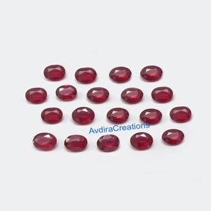 May include: A collection of oval-shaped, deep red gemstones. The gems are evenly spaced against a white background. The text "AvdiraCreations" is visible in the image. These gemstones are suitable for jewelry making.
